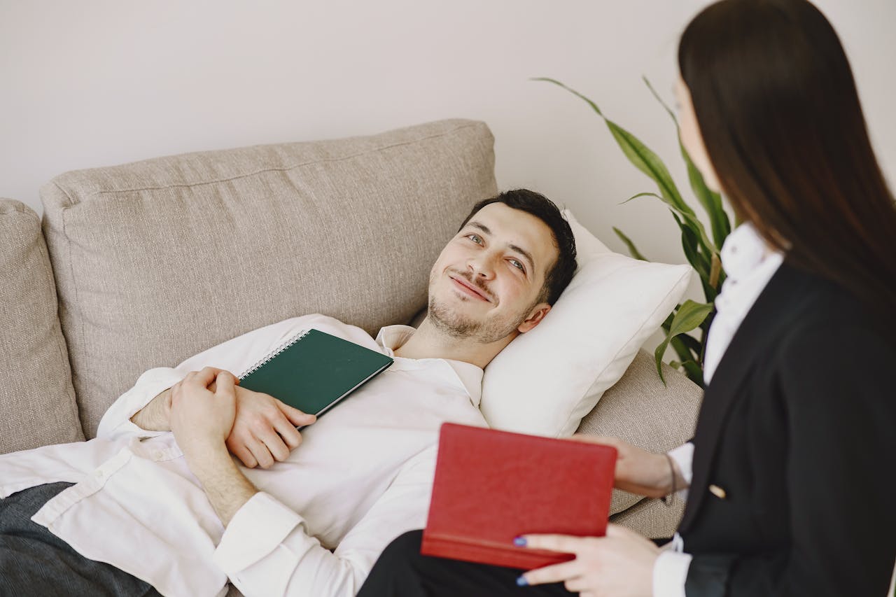 Positive man in formal wear lying on sofa with notebook in hands while smiling and talking with therapist in black suit